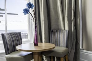 A round table with two upholstered chairs, a vase with flowers, and a view of the sea through a window with curtains. - Majestic Hotel Eastbourne by Belvilla - single sea view (Eastbourne)