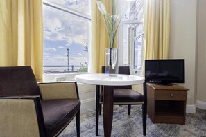 A hotel room with a round table, a TV on a wooden stand, a chair, a vase with plants, and a window with a view of the sea.