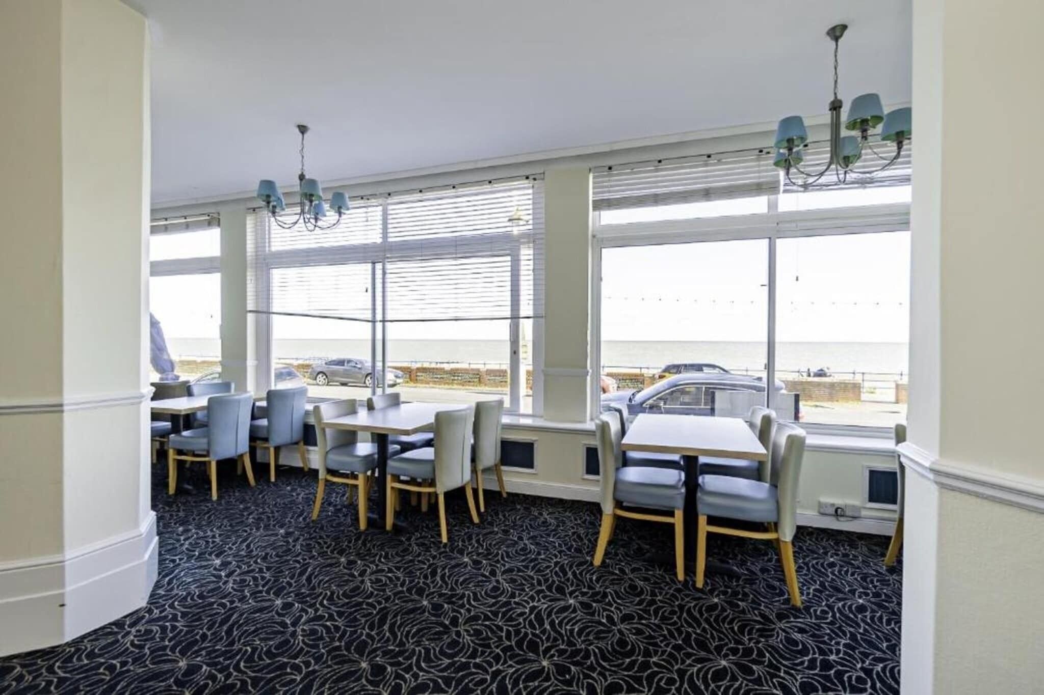 Ein Essbereich mit Tischen und Stühlen, großen Fenstern mit Jalousien und Blick auf das Meer.