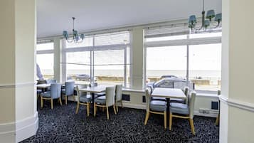 Ein Essbereich mit Tischen und Stühlen, großen Fenstern mit Jalousien und Blick auf das Meer.
