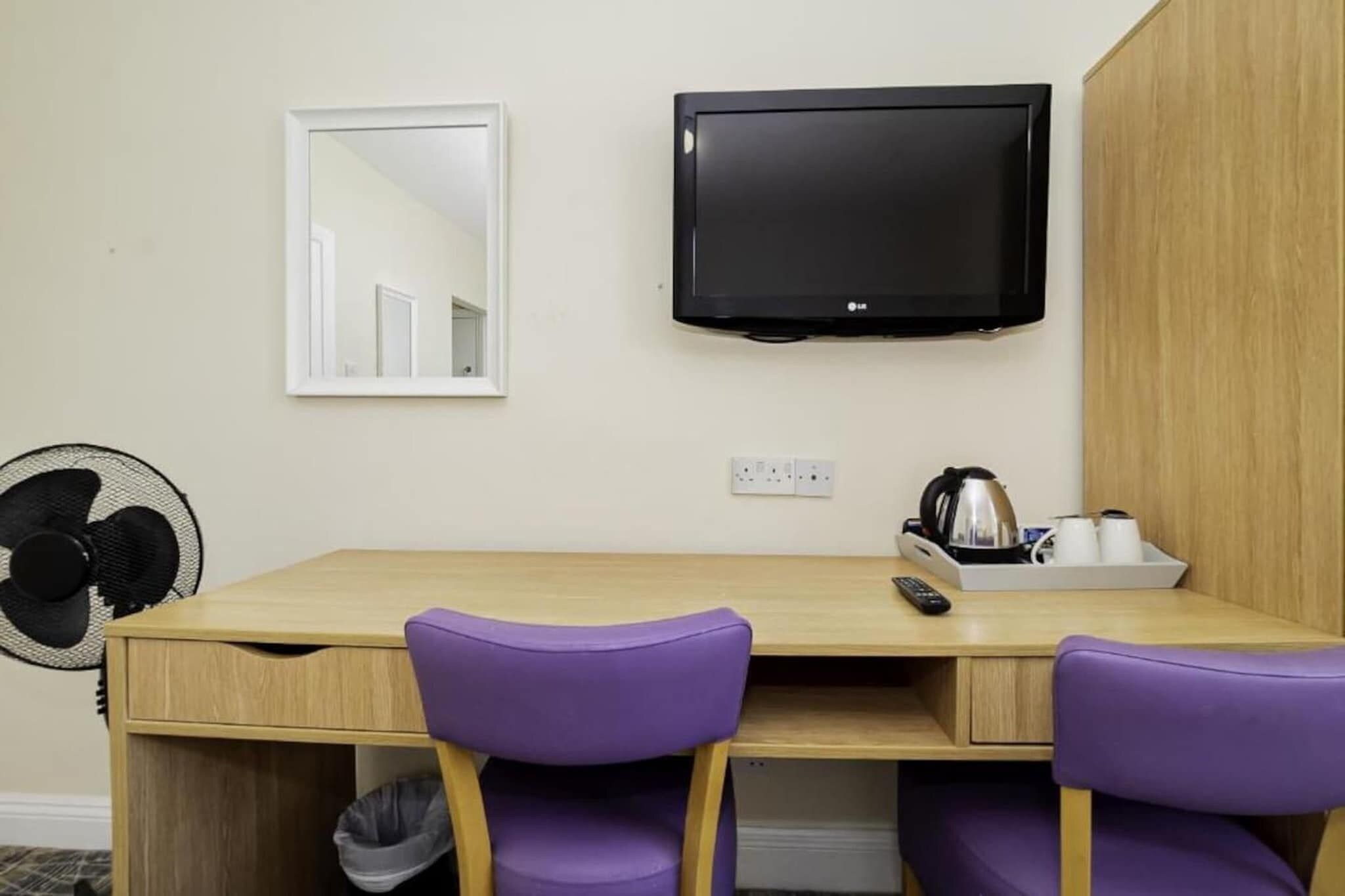 Une pièce avec un bureau en bois, deux chaises violettes, un téléviseur noir fixé au mur, un miroir, un ventilateur et une bouilloire posée sur le bureau.