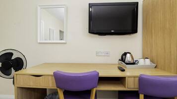 Une pièce avec un bureau en bois, deux chaises violettes, un téléviseur noir fixé au mur, un miroir, un ventilateur et une bouilloire posée sur le bureau.