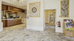 Un área de recepción con piso de mármol, un candelabro, un mostrador de recepción, un reloj y un extintor.