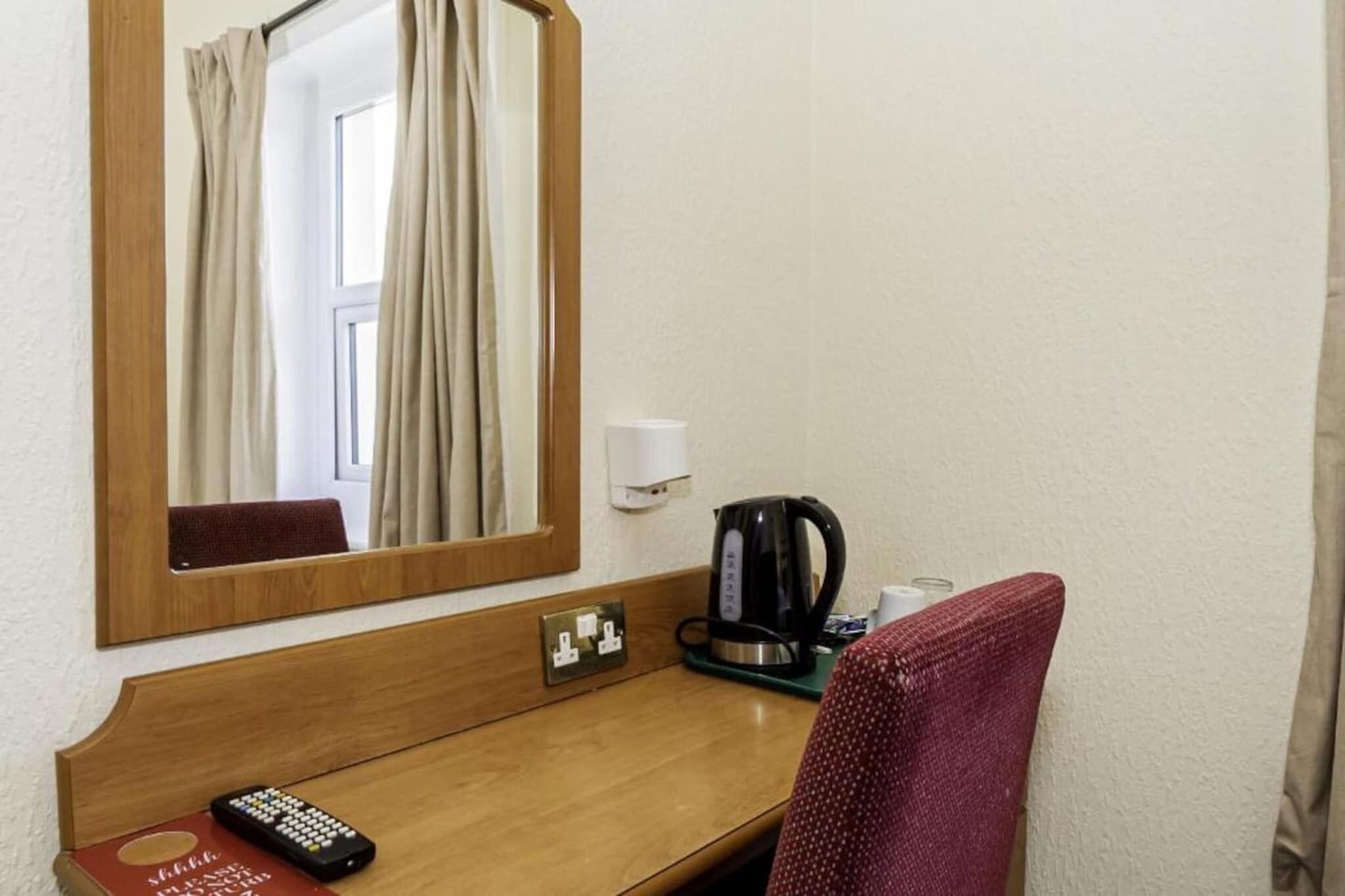 Habitación de hotel con escritorio de madera, espejo, cafetera, control remoto y una silla roja.
