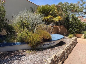 A boat-shaped planter with the name 'Hacienda' on a patio with gravel, surrounded by various plants and trees.