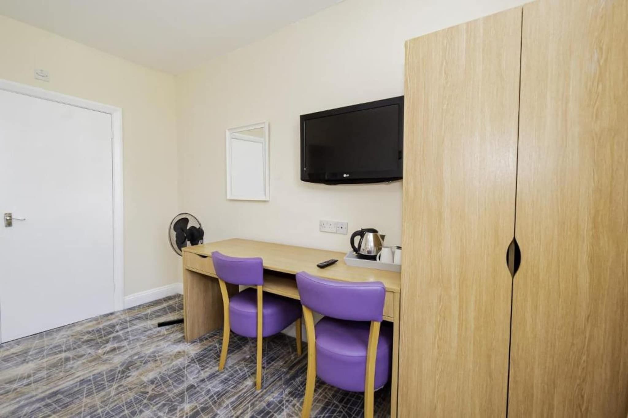 Une pièce comprenant une armoire en bois, un téléviseur fixé au mur, un bureau avec deux chaises violettes et une machine à café.
