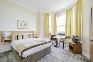 A hotel room with a bed, bedside table, fan, TV, and a view of the sea. - Double room with sea view (Eastbourne)