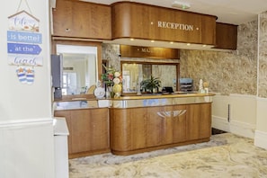 Uma área de recepção com balcão de madeira, uma placa com a inscrição 'RECEPTION' e um espelho acima.