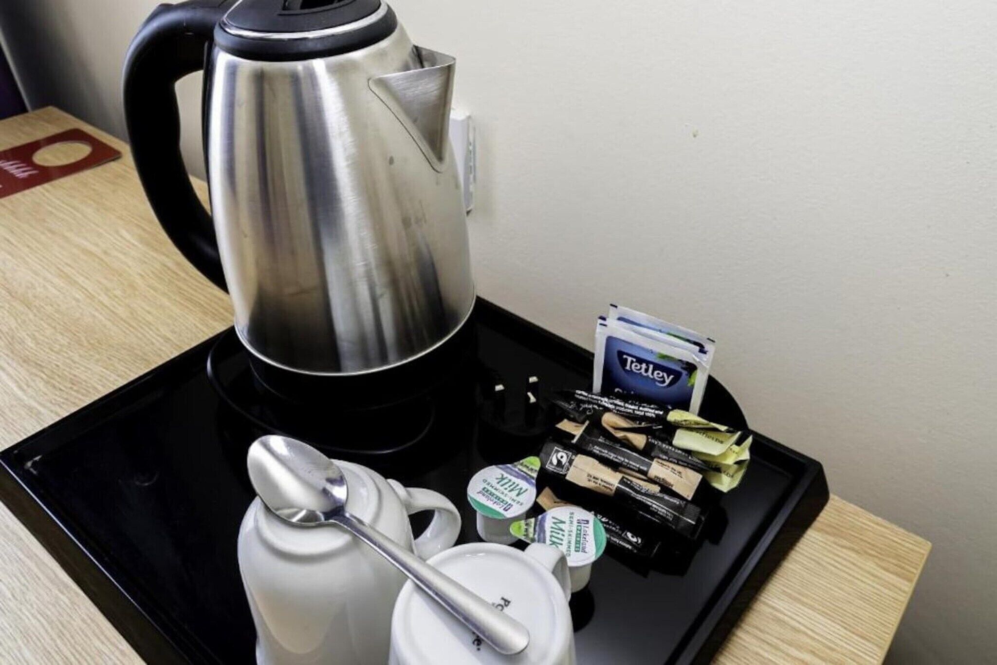 A stainless steel kettle, a white teapot, a tray with Tetley tea bags, and a milk carton on a black tray.