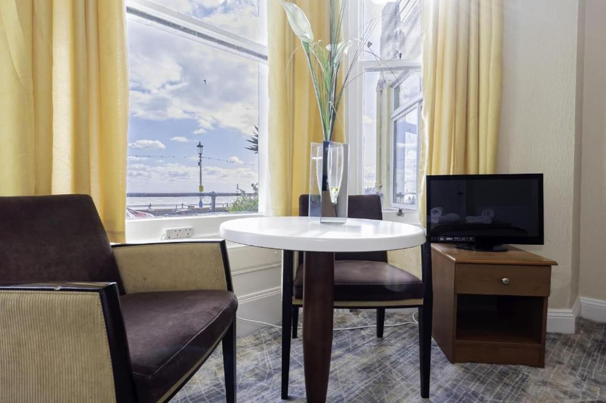 A hotel room with a round table, a television on a wooden stand, a chair, and a view of the sea through large windows.