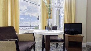 Habitación de hotel con una mesa redonda, un televisor sobre un soporte de madera, una silla y vista al mar a través de amplios ventanales.