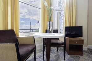 Une chambre d’hôtel avec une table ronde, un téléviseur posé sur un meuble en bois, une chaise et une vue sur la mer par de grandes fenêtres.