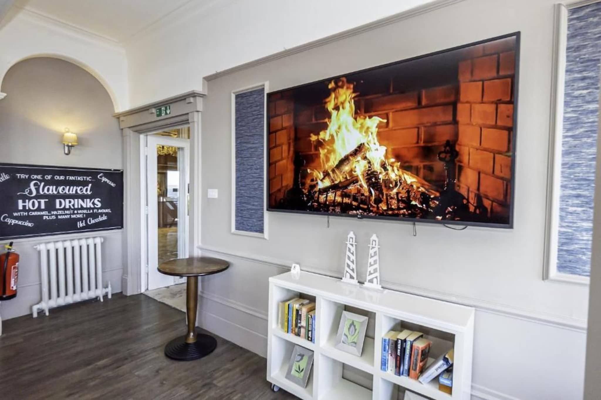 A hotel lobby with a fireplace, a sign for hot drinks, and a bookshelf.
