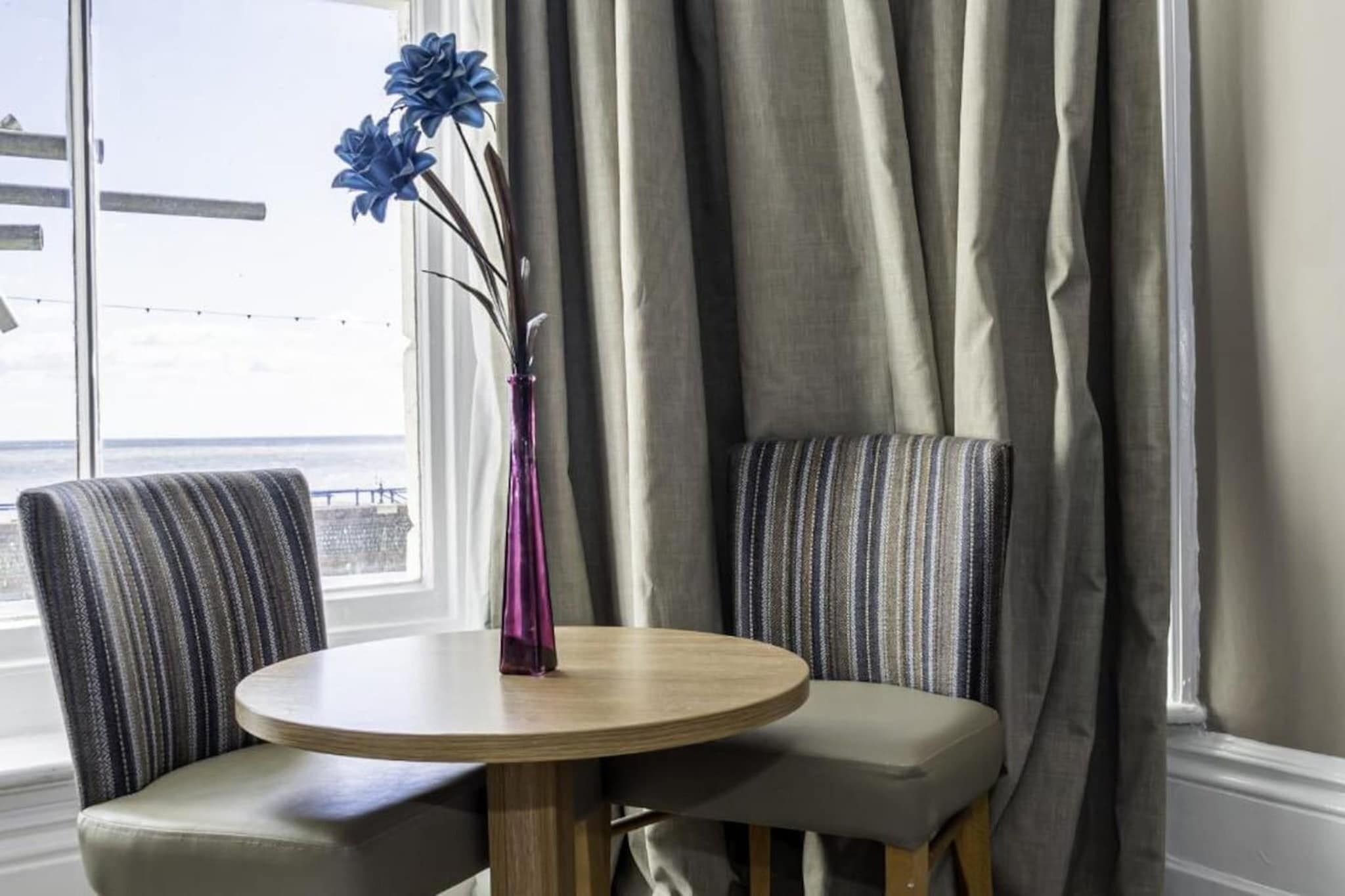 A room with a round table, two striped chairs, a vase with blue flowers, and a view through a window with curtains.