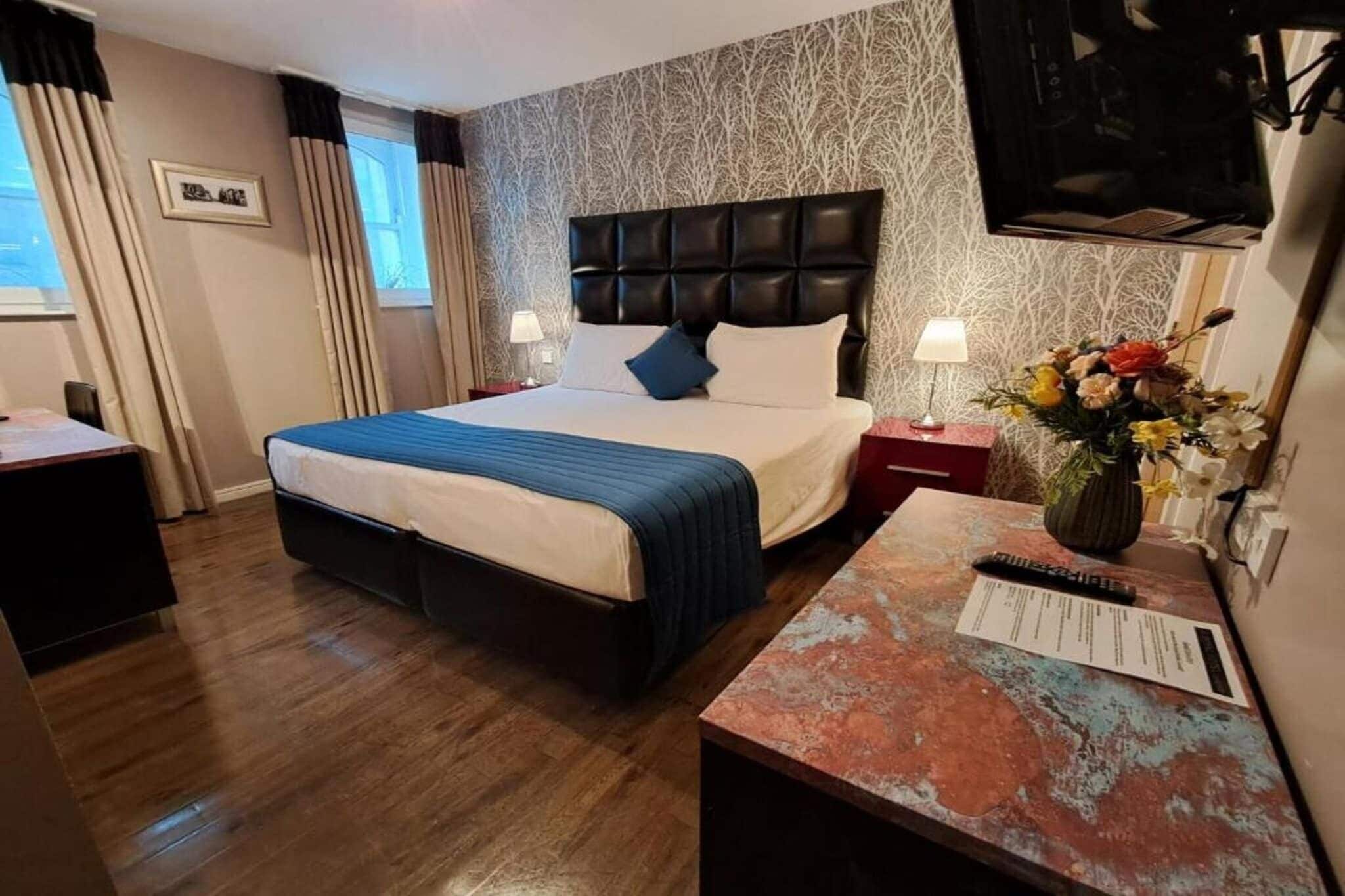 A hotel room with a bed, two bedside tables, a TV, and a desk with a vase of flowers.