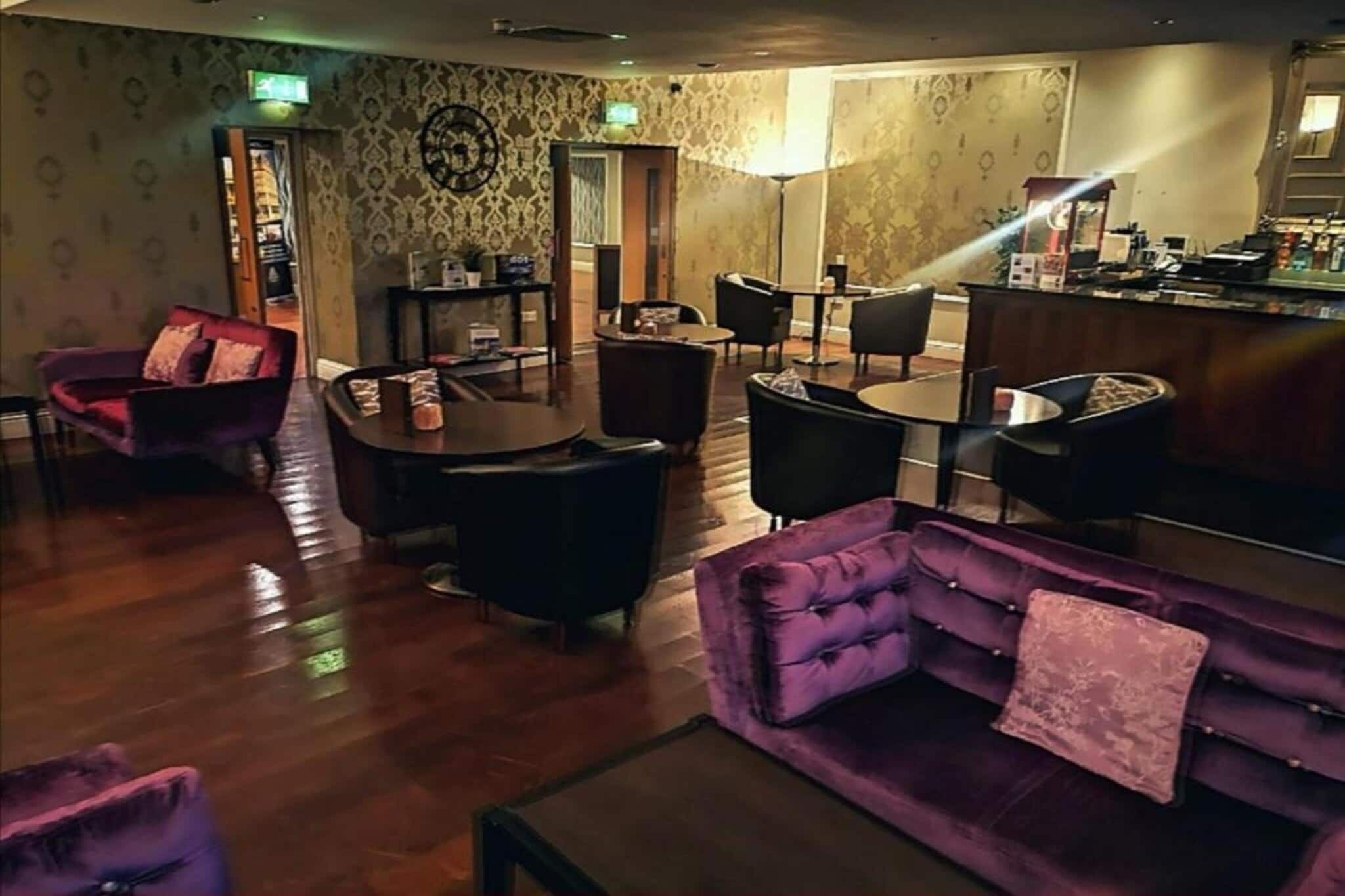 Un hall d’hôtel avec un canapé violet, des chaises noires, un plancher en bois et un papier peint à motifs.