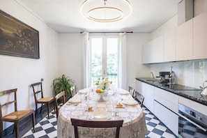 Un coin repas avec une table dressĂ©e pour quatre personnes, une cuisine Ă©quipĂ©e dâappareils modernes et une grande fenĂȘtre avec des rideaux.