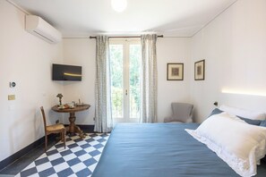 A hotel room with a checkered floor, a bed with blue bedding, a television mounted on the wall, a small table with a lamp, and a chair.