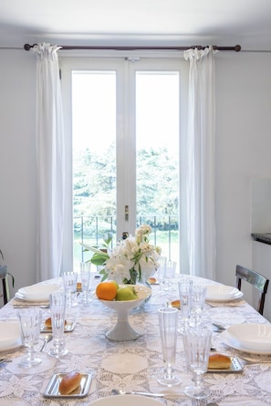 Une table Ă manger dressĂ©e avec des assiettes, des verres et un saladier de fruits, avec vue sur les arbres Ă travers une fenĂȘtre.