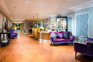 Eine Hotelhalle mit Rezeption, violetten Sofas, Holzböden und einer Wand mit Muster.