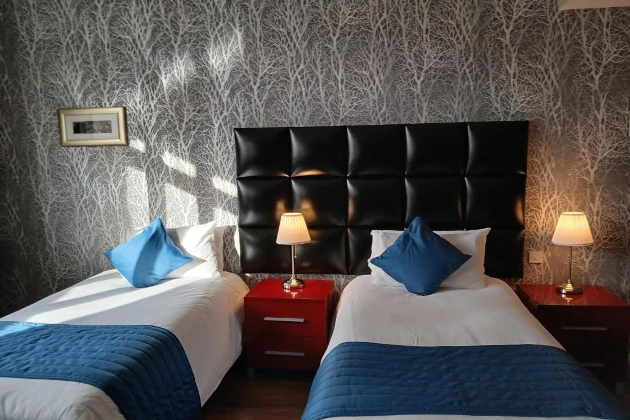 A hotel room with two beds, a dark headboard, bedside lamps, and a patterned wallpaper.
