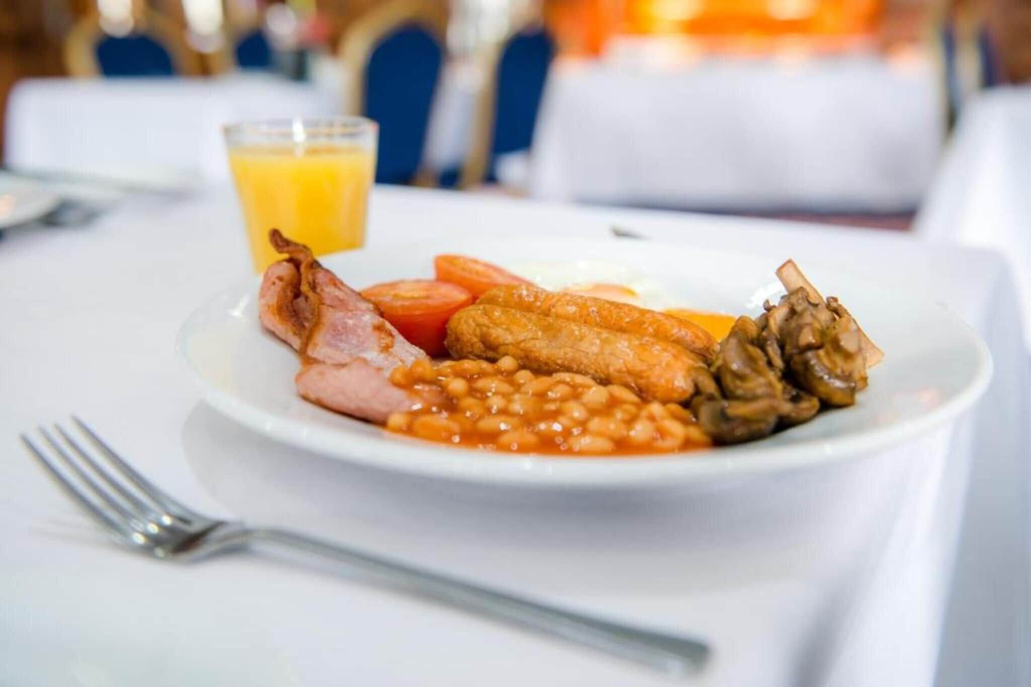A plate of breakfast food including bacon, beans, sausages, and mushrooms, with a glass of orange juice on the side.