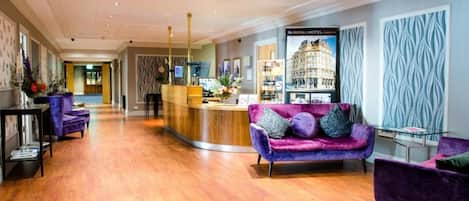 A hotel lobby with a reception desk, purple sofas, wooden flooring, and a patterned wall.