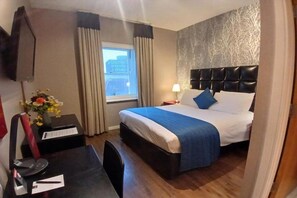 A hotel room with a bed, a desk, a chair, a lamp, a window with curtains, and a wall with a tree pattern. - Standard Double Room - No Window (Cardiff)