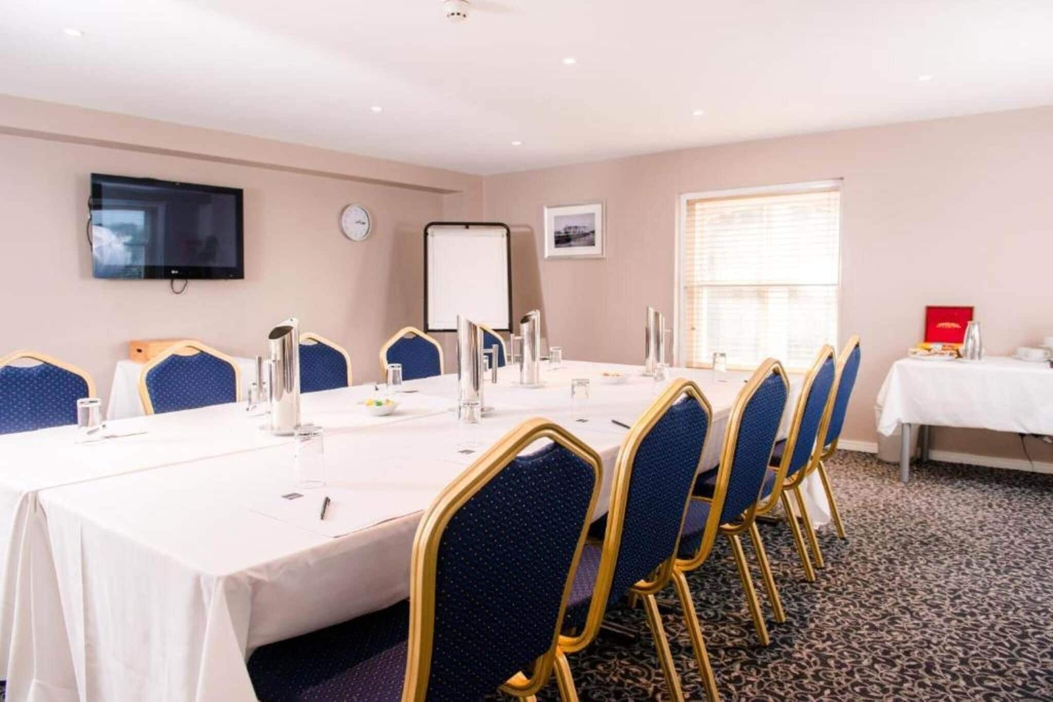 A conference room with a long table, chairs, a whiteboard, a clock, and a window with blinds.