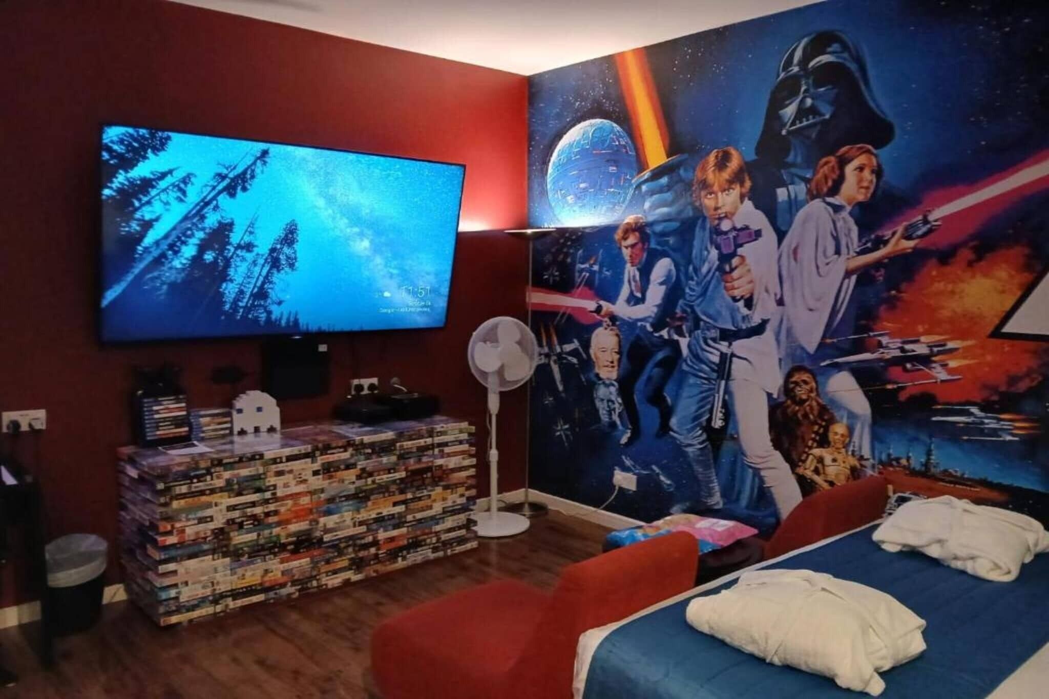 A room with a large TV, a bookshelf, a bed, and a wall mural.