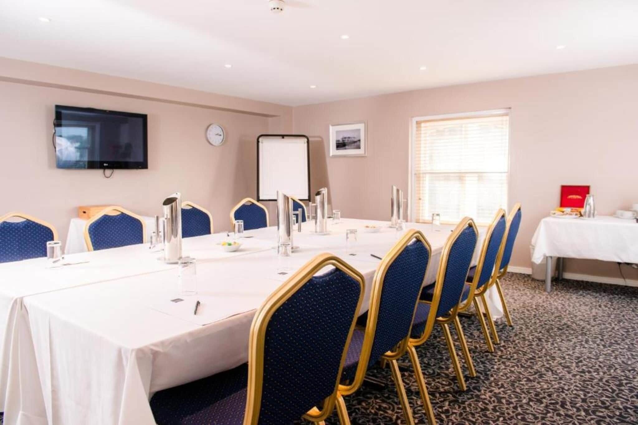 A conference room with a long table, chairs, a whiteboard, a clock, and a window with blinds.