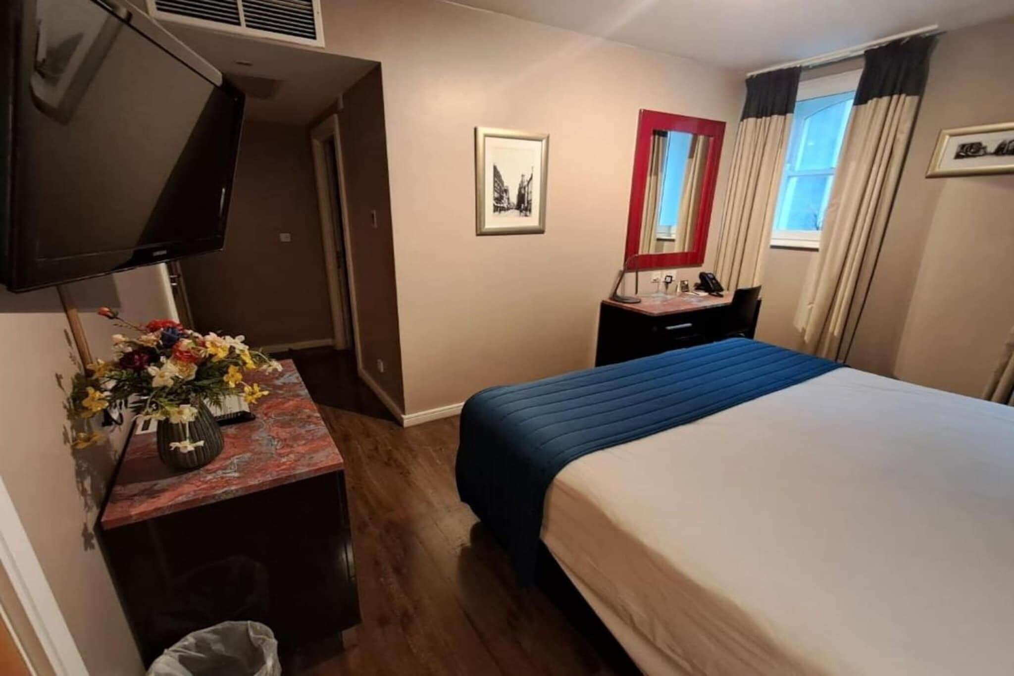 Ett hotellrum med en säng, ett skrivbord med en vas med blommor, en tv och ett fönster med gardiner.