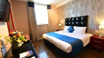 Ein Hotelzimmer mit einem Bett, einem Nachttisch, einem Fernseher und einem Schreibtisch mit Blumen in einer Vase.