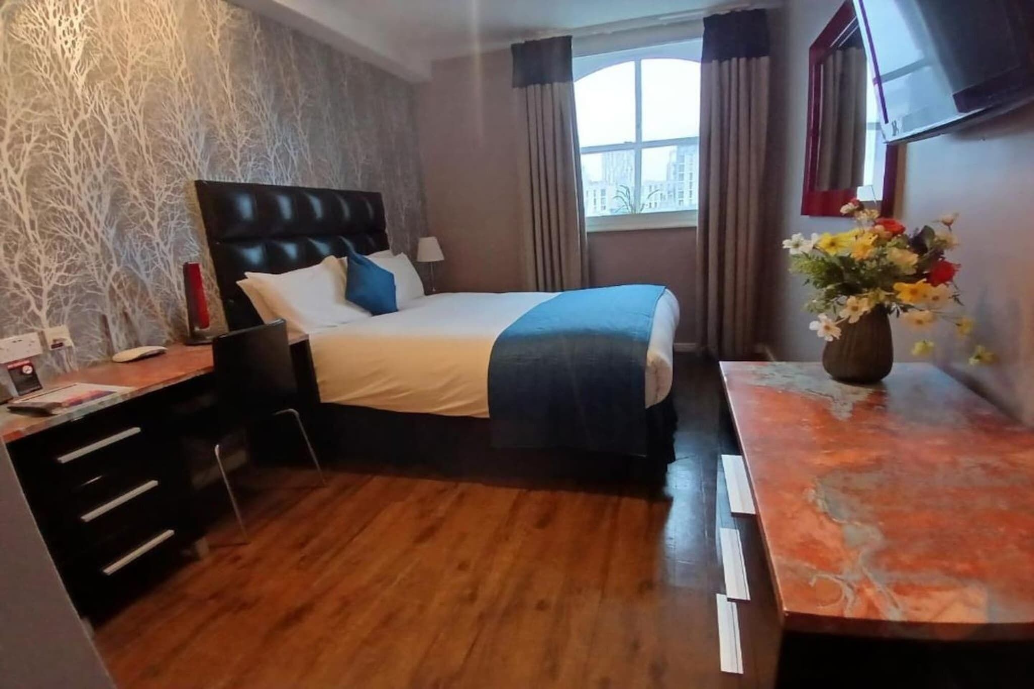 A hotel room with a bed, a desk, a chair, a window with curtains, and a vase with flowers.