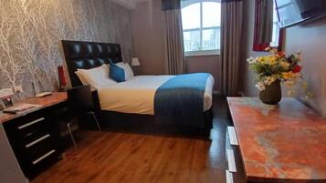 A hotel room with a bed, a desk, a chair, a window with curtains, and a vase with flowers.