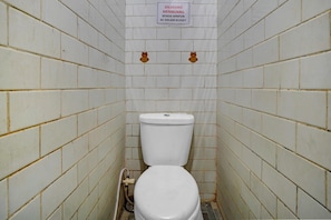 Une petite salle de bain carrelée, avec des toilettes, une pancarte au-dessus et un tuyau sur le sol.