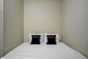 Une petite chambre avec un lit simple, dotée de draps blancs et de deux oreillers noirs ornés d’une bande marron.