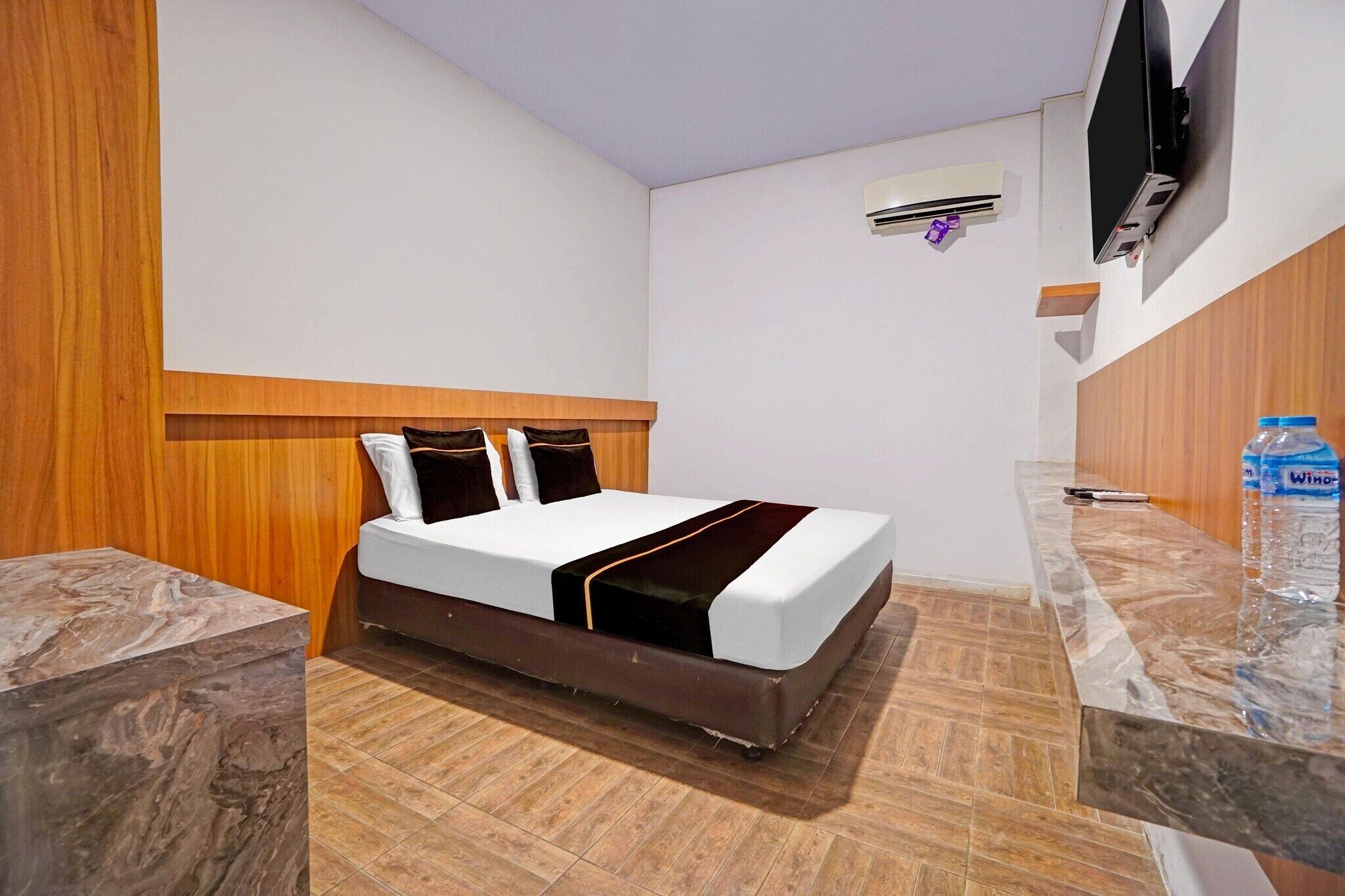 Camera d'albergo con un letto, un comodino in marmo, una testiera in legno, una TV a schermo piatto fissata al muro e una bottiglia d'acqua sul ripiano.