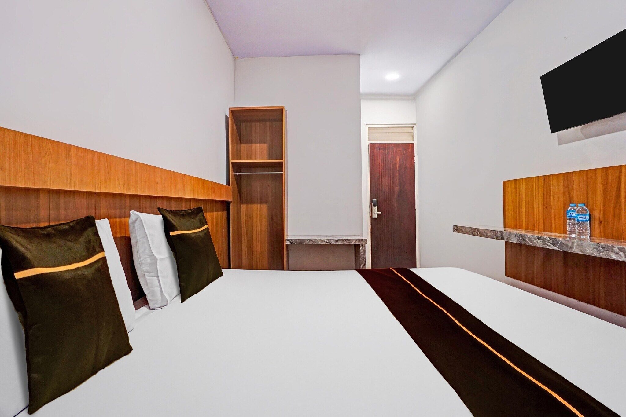 Quarto de hotel com cama, cabeceira de madeira, pequena escrivaninha, espelho e televisão fixada na parede.