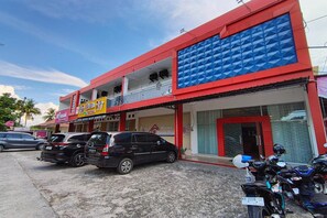 Un bâtiment à la façade rouge et bleue, avec une enseigne bien visible indiquant « Wisma Esun Budi », et des voitures et des motos garées devant.