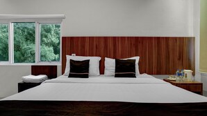 Quarto de hotel com uma cama grande, cabeceira de madeira, mesa de cabeceira com abajur e janela com vista para árvores.