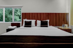 A hotel room with a large bed, a wooden headboard, a nightstand with a lamp, and a window with a view of trees.