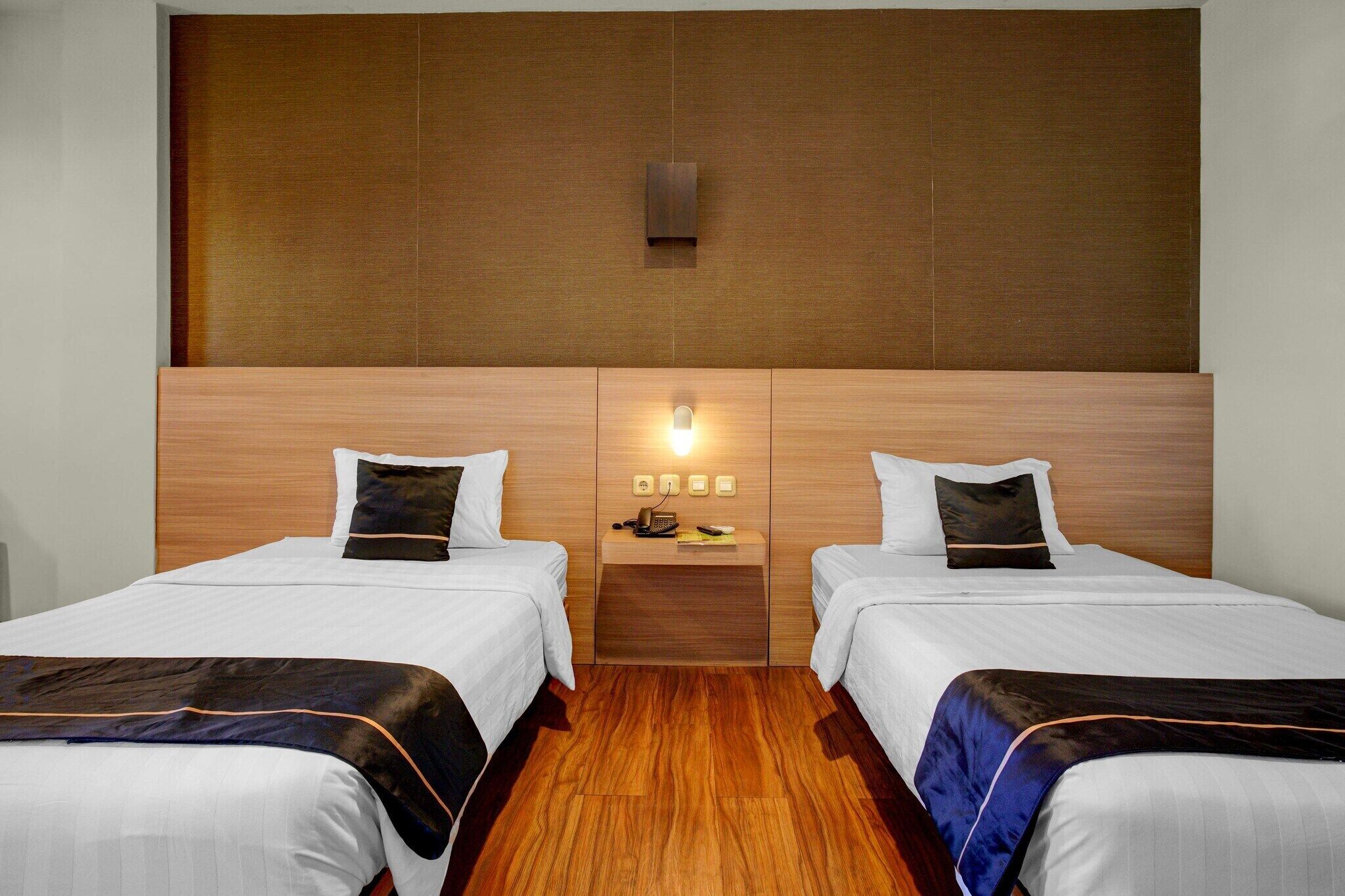Une chambre d’hôtel avec deux lits, une tête de lit en bois, une table de chevet avec une lampe et une petite étagère avec un téléphone et un livre.