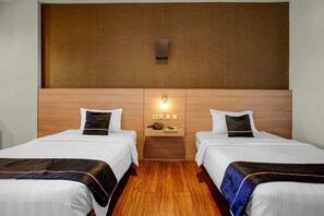 A hotel room with two beds, a wooden headboard, a bedside table with a lamp, and a small shelf with a phone and a book.