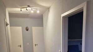 Un corridoio con pavimento in legno, pareti bianche e una zona cucina con pensili e un piano di lavoro visibile attraverso una porta aperta.