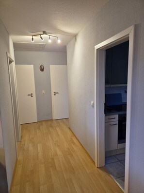 Un passadís amb sòl de fusta, parets blanques, i una cuina amb armaris i una superfície de treball visibles a través d'una porta oberta.