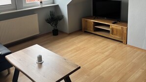 Un soggiorno moderno con pavimento in legno, una TV su un mobile in legno, un termosifone, una finestra con vista su un edificio in mattoni e una piccola pianta.