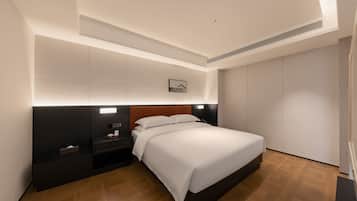 A modern hotel room with a large bed, a nightstand, a small table, and a wardrobe.