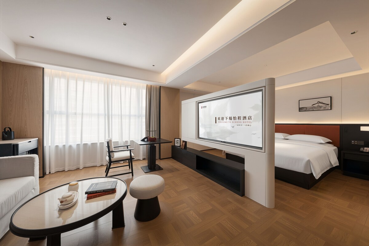 Une chambre d’hôtel moderne équipée d’un lit, d’un bureau, d’une chaise, d’une table ronde et d’un écran de télévision affichant un message de bienvenue.
