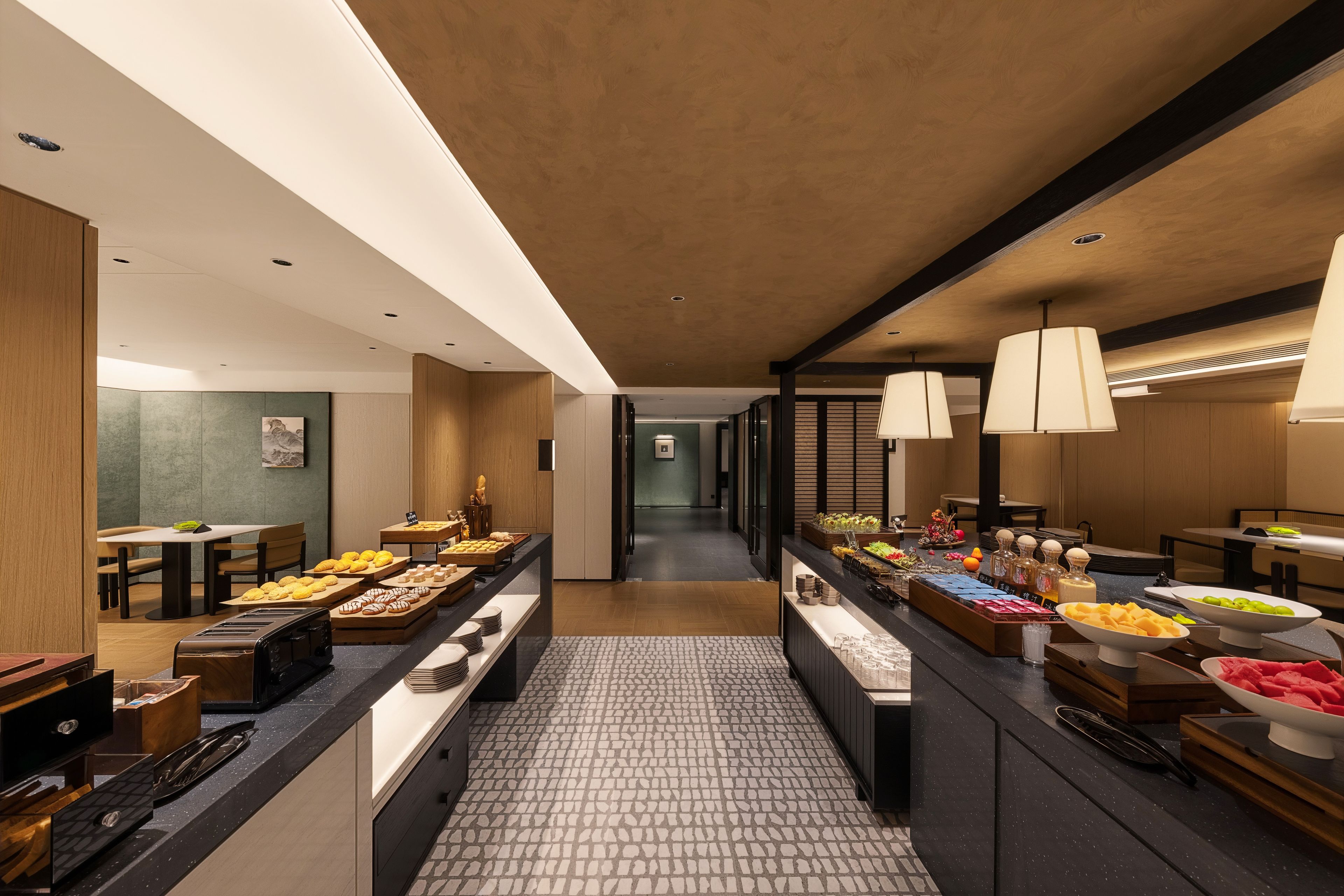 A modern hotel lobby with a central food display, seating areas, and a view into the corridor.